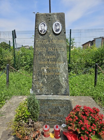 Cippo commemorativo dedicato a Daccò e Polli, circondato da fiori e candele in un contesto verde e urbano.