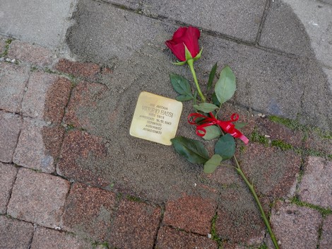 Pietra d'inciampo dedicata a Bassi, adornata con una rosa rossa e un nastro rosso, su un pavimento di ciottoli.