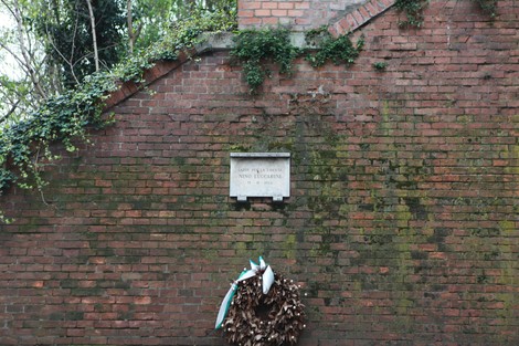 Lapide commemorativa su un muro di mattoni, decorata con una corona di foglie e un nastro.