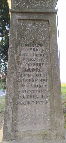 Monumento ai caduti dell'Amola con iscrizioni commemorative e circondato da un paesaggio rurale.