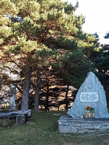 Monumento commemorativo con dettagli in pietra e decorazioni floreali, circondato da alberi.