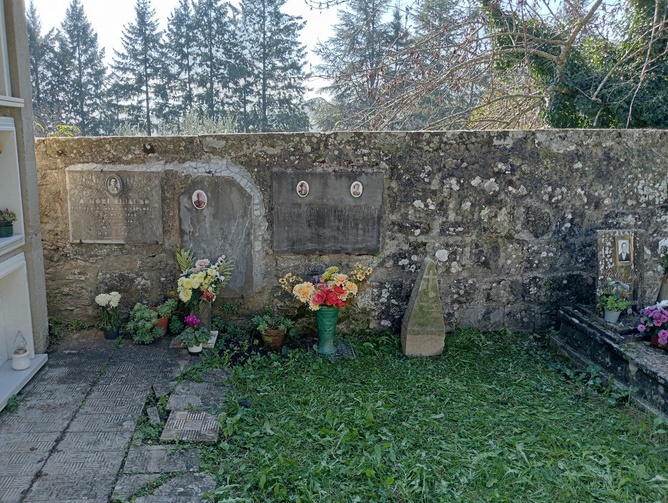 Cippo a Videschi con tombe e fiori, circondato da un muro di pietra e alberi sullo sfondo.