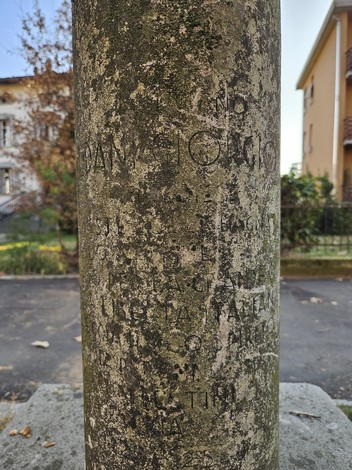 Cippo in pietra con iscrizioni, circondato da vegetazione e edifici, simbolo di memoria storica.