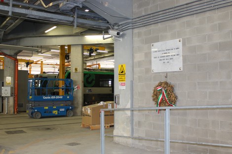 Lapide commemorativa ad Avezzano con una corona di fiori, in un ambiente industriale con veicoli e attrezzature.