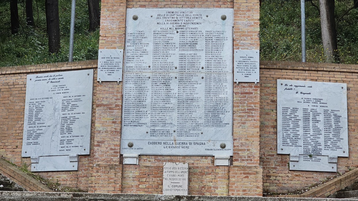 Monumento ai caduti con targa commemorativa e nomi dei caduti in guerra, incorniciato da bosco.