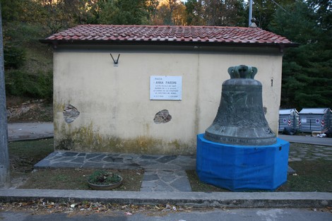 Lapide commemorativa di Annal Pardini con una campana all'esterno, situata in un contesto naturale.