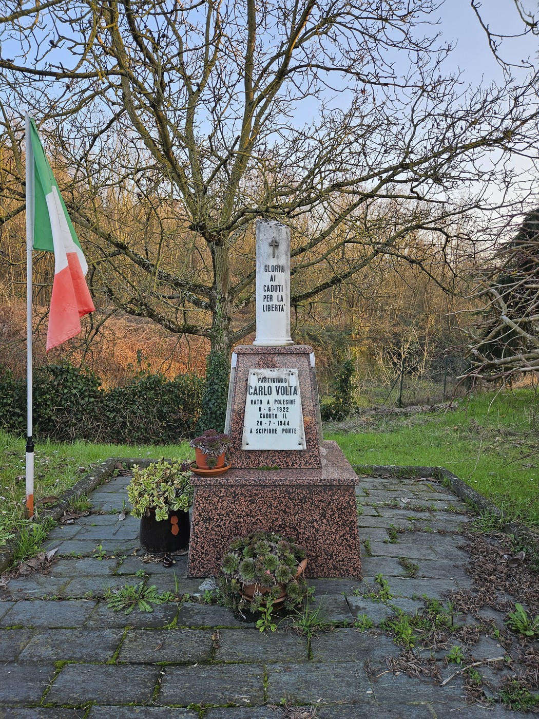 Cippo commemorativo circondato da vegetazione, con bandiera italiana e piante ornamentali.