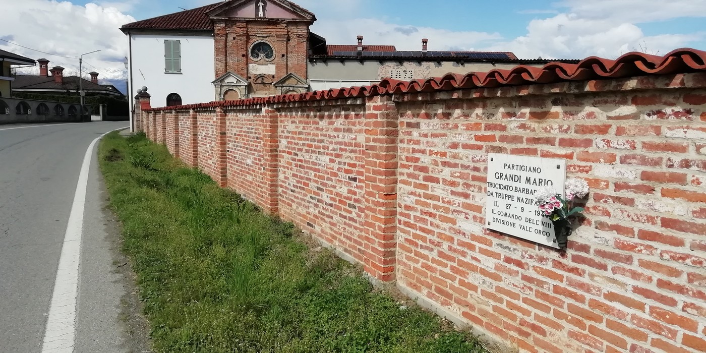 Lapide commemorativa a Grandi, con muro di mattoni e campagna circostante. Scena di quiete e riflessione.