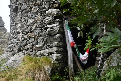 Rifugio partigiano in pietra circondato da vegetazione, con bandiera appesa all'ingresso.