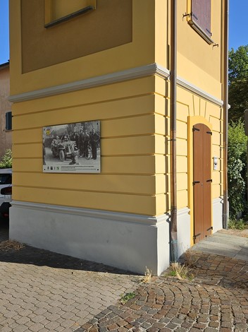 Targa commemorativa su un edificio giallo, con immagine storica e gruppi di persone in primo piano.