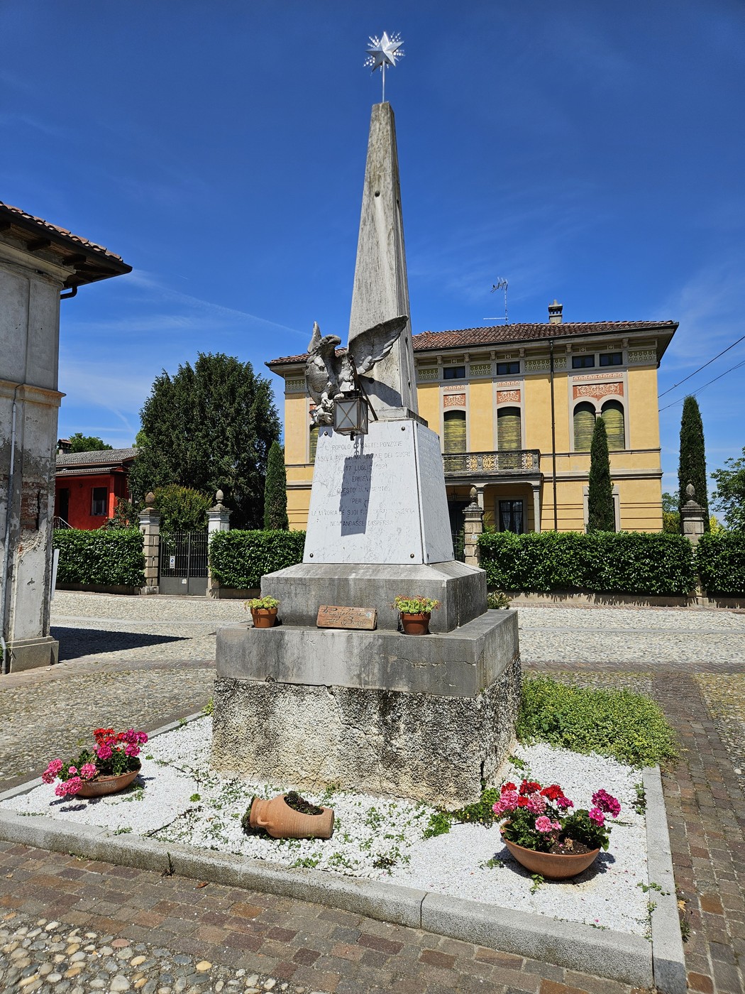 Monumento ai Caduti di Castelponzone, con obelisco e decorazioni, circondato da piante e fiori.