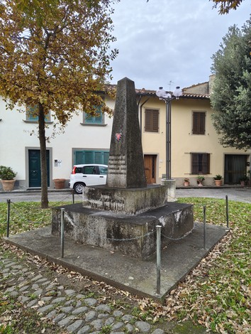 Monumento ai deportati, obelisco di pietra circondato da alberi e case, in una piazza tranquilla.