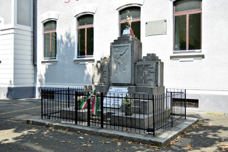 Monumento in pietra con bassorilievi e iscrizioni, recintato da cancellata, davanti a un edificio.