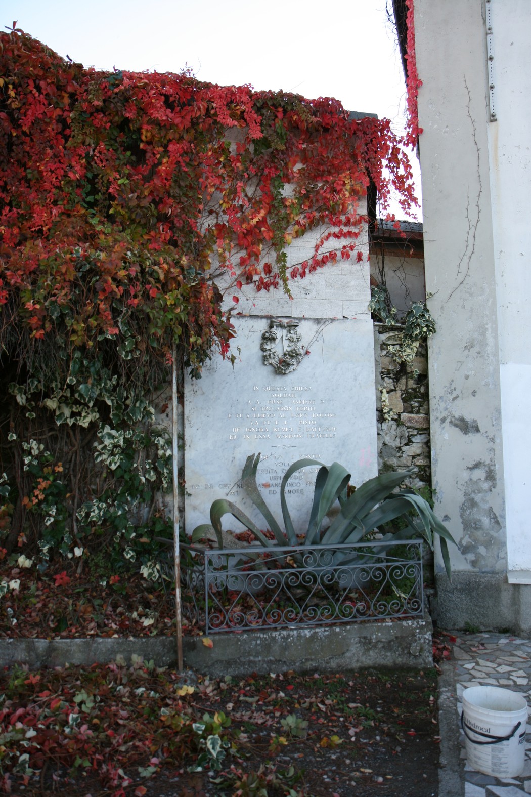 Lapide della chiesa di Santa Lucia, adornata da edera rossa e circondata da una vegetazione rigogliosa.
