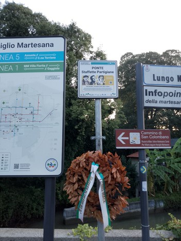 Targa commemorativa del Ponte Staffette Partigiane con corona di alloro, circondata dalla vegetazione.