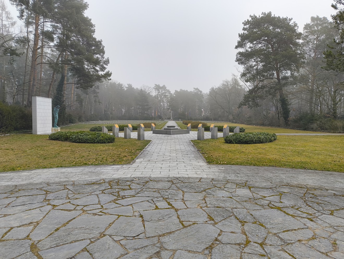 Cimitero militare italiano di Zehlendorf con un percorso centrale e lapidi in un ambiente boschivo nebbioso.