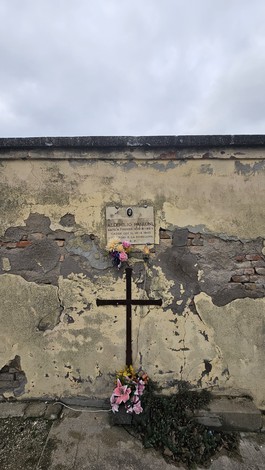 Croce in legno posta su un muro screpolato, adornata con fiori e una targa commemorativa.
