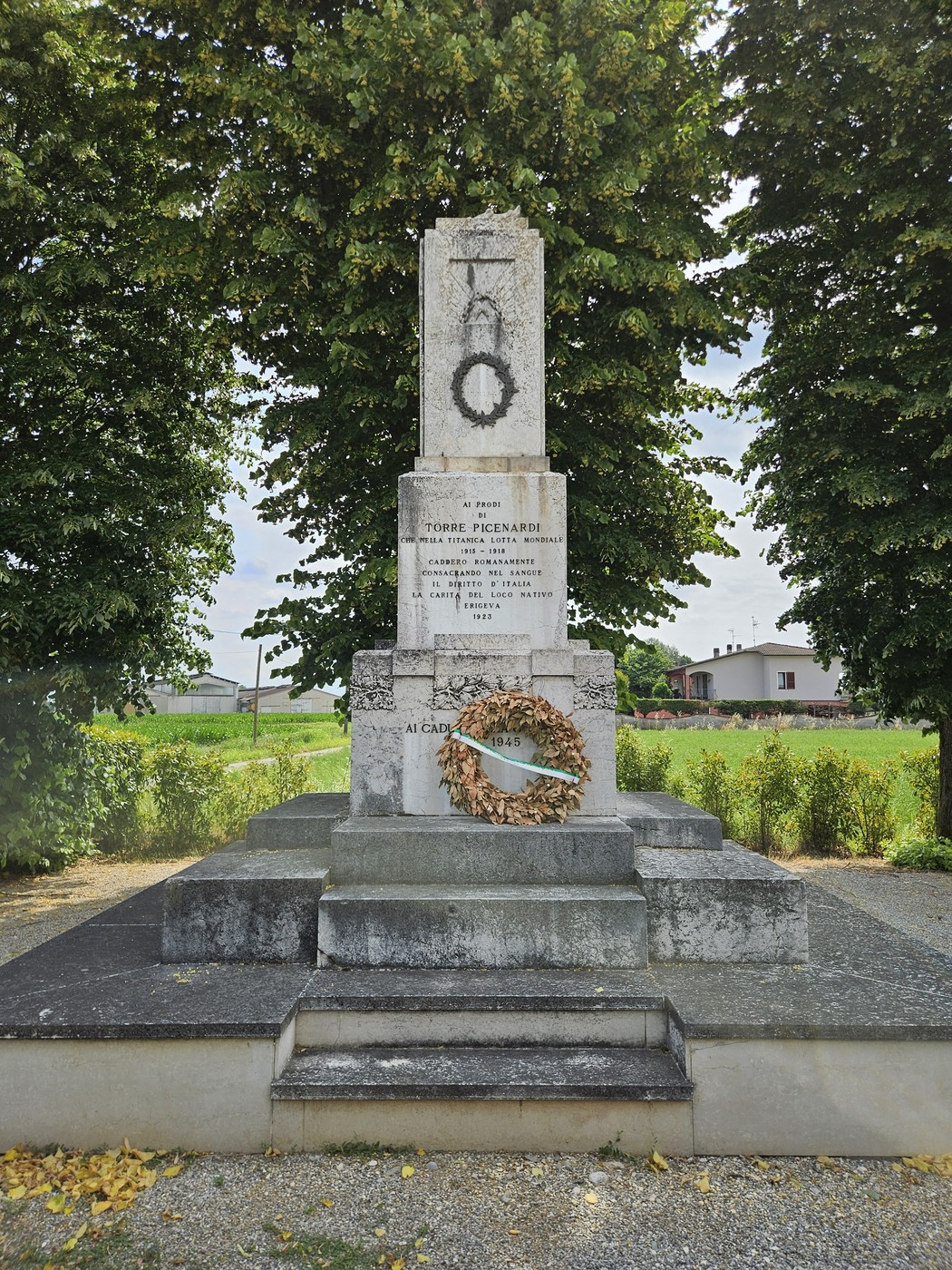 Monumento ai Caduti con una corona d'alloro, circondato da alberi e un paesaggio rurale sullo sfondo.