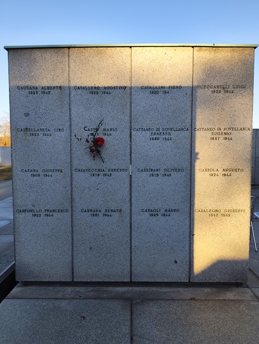 Monumento commemorativo al Campo della Gloria con nomi incisi, fiori e un'atmosfera di rispetto e memoria.