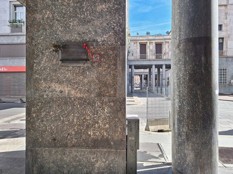 Lapide commemorativa su una colonna, affiancata da elementi architettonici e spazi urbani di Martorelli.