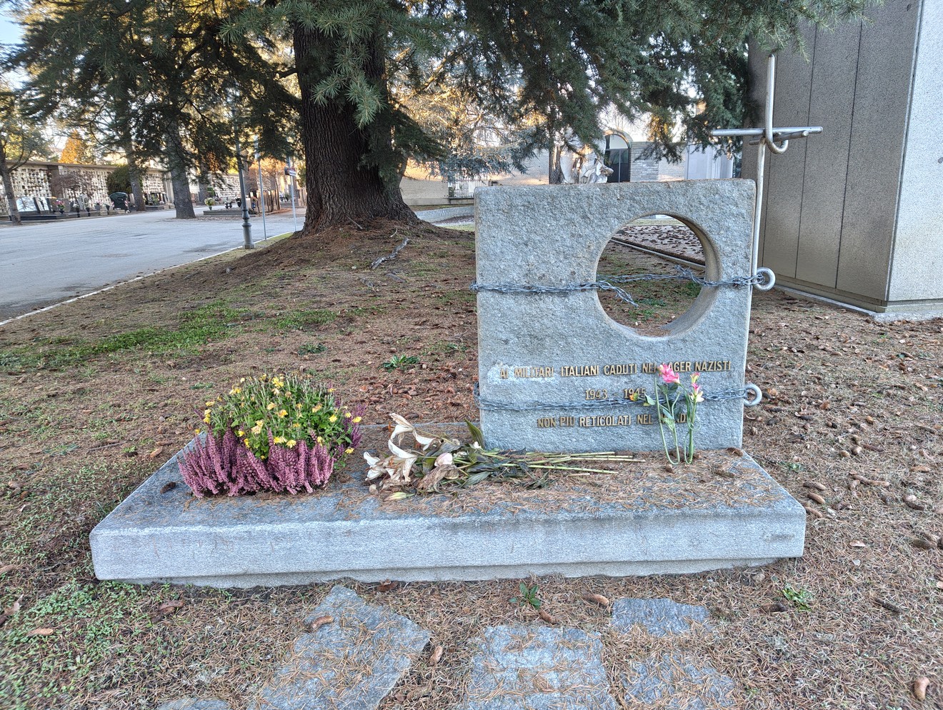 Monumento agli Internati Militari Italiani, con fiori e decorazioni su una base in pietra, circondato da alberi.