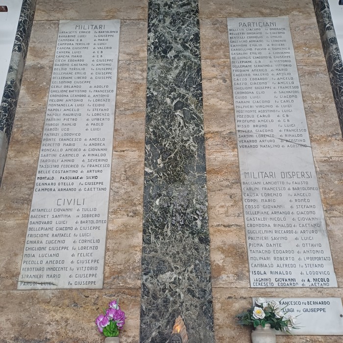 Due lapidi marmoree affiancate con lunghi elenchi di nomi, sotto fiori e una lampada votiva.