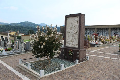 Monumento ai Caduti partigiani in un cimitero, circondato da alberi e tombe, con montagne sullo sfondo.