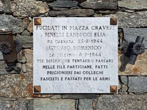 Lapide marmorea con iscrizione, fissata a un muro in pietra, affiancata da due elementi in ferro battuto.