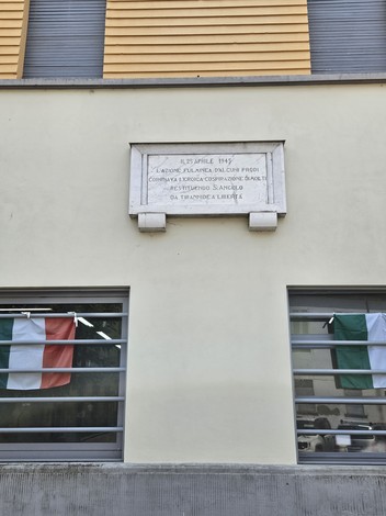 Lapide commemorativa della Liberazione di Sant'Angelo, situata su un edificio, con bandiere italiane nelle vicinanze.