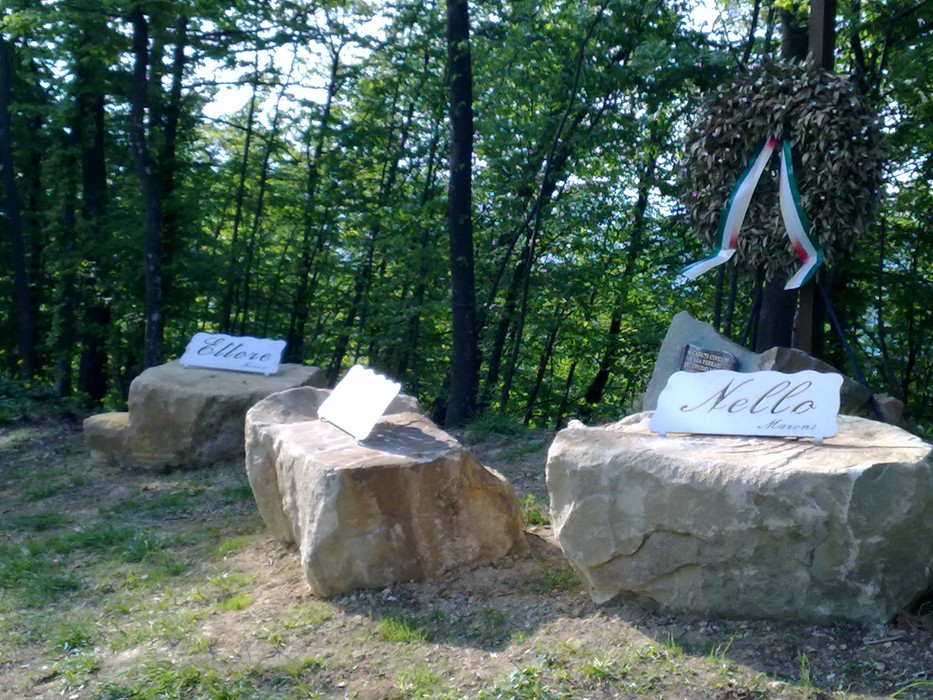 Monumento commemorativo ai Ferrari e a Maroni, collocato in un contesto naturale tra alberi e rocce.