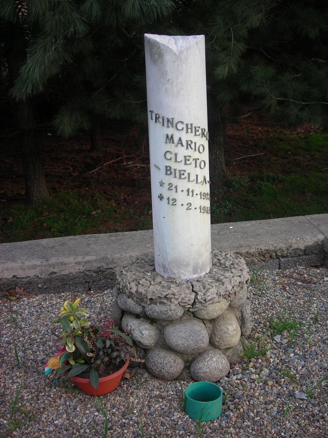 Cippo commemorativo dedicato a Mario Cleto Trinchero, con fiori e pietre decorative attorno.