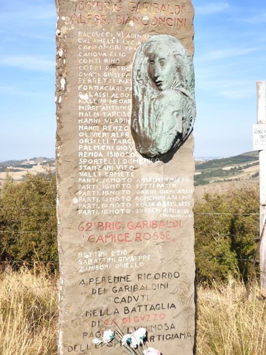 Stele in pietra con iscrizioni e un rilievo a forma di volto, collocata in un prato con il paesaggio collinare sullo sfondo.