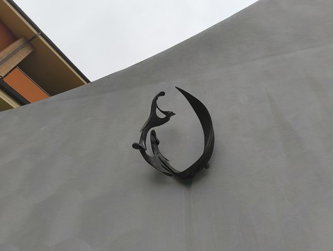 Monumento a Piccinini, scultura in metallo appoggiata su una parete grigia, con forma evocativa e sinuosa.