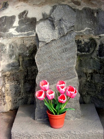 Cippo commemorativo a Mentegazzi con fiori rosa in un vaso, incastonato in una parete di pietra.