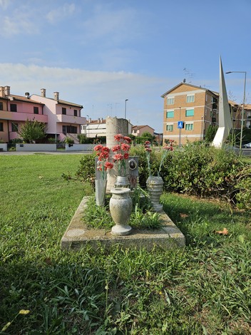 Cippo commemorativo con fiori e decorazioni, situato in un'area verde urbana. Edifici sullo sfondo.