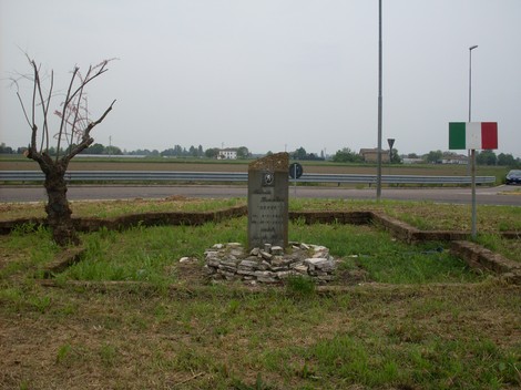 Cippo commemorativo immerso nel verde, con strada e paesaggio al fondo. Atmosfera di riflessione e memoria.