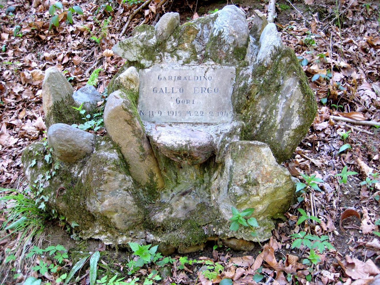 Cippo commemorativo in pietra situato nel bosco, circondato da rocce e vegetazione.