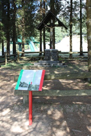 Crocifisso dei Partigiani immerso nel verde, con panchine in legno e pannello informativo di fronte.