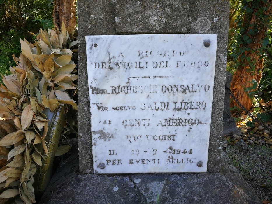 Lapide in marmo con iscrizione, fissata su un cippo in pietra, tra la vegetazione.