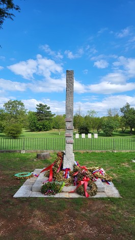Monumento in pietra a forma di stele, con targhe incise, circondato da corone di fiori nel prato.