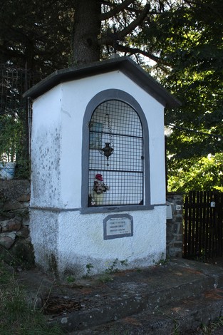 Lapide commemorativa in un edicolo, con fiori e decorazioni, immersa nel verde della natura circostante.