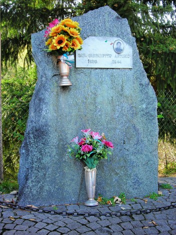 Monumento a Gasparetto, con lapide commemorativa e fiori, situato in un ambiente naturale.