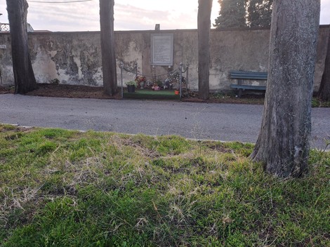 Lapide in un'area verde, circondata da alberi e fiori, con una panchina sullo sfondo.