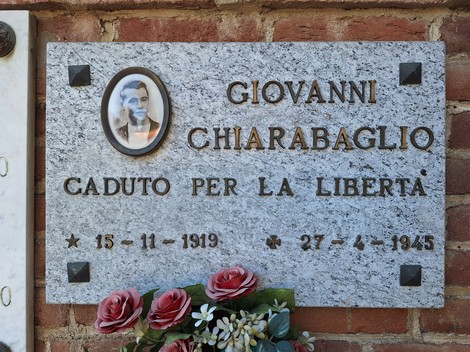 Lapide in granito con foto ovale e iscrizione, fissata a muro di mattoni, con fiori appoggiati sotto.