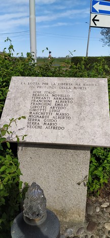 Stele commemorativa ai caduti, incisa su pietra, circondata da vegetazione e in prossimità di una strada.