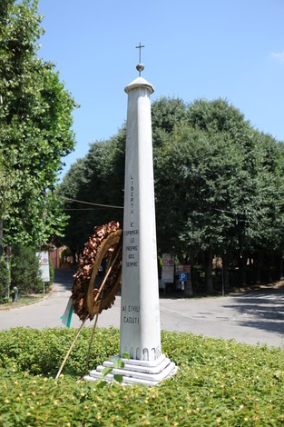 Monumento ai caduti circondato da vegetazione, con una corona di fiori e il cielo blu sullo sfondo.