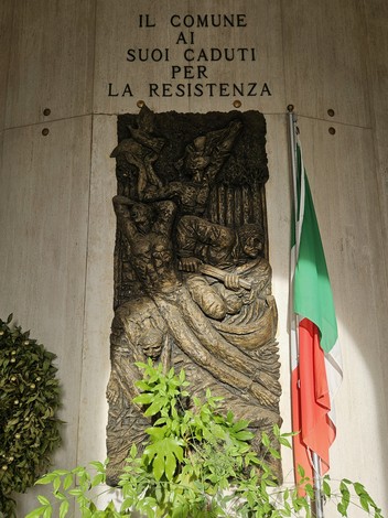 Scultura commemorativa al "Sacrario dei Caduti per la Resistenza" con bandiera italiana e vegetazione circostante.