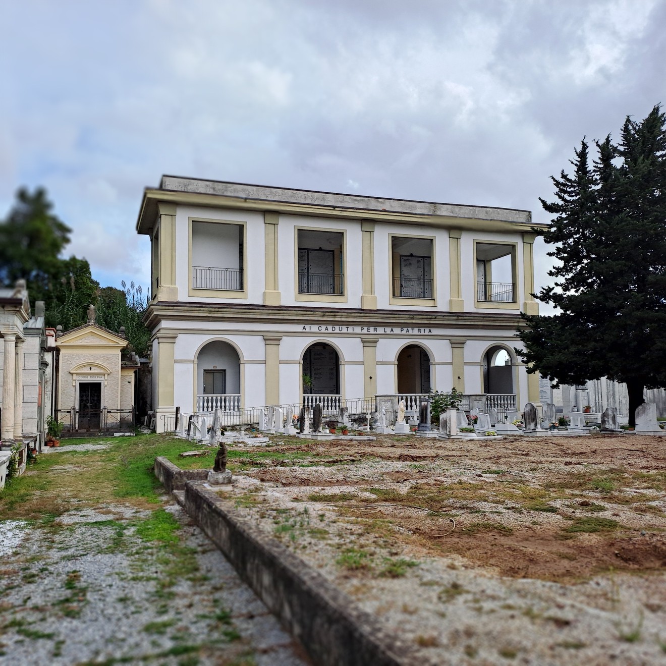 Edificio a due piani con portico ad arcate e iscrizione, circondato da lapidi in un cimitero.