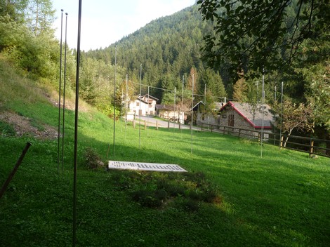 Monumento ai Dodici Caduti immerso in un paesaggio verde, circondato da alberi e montagne.