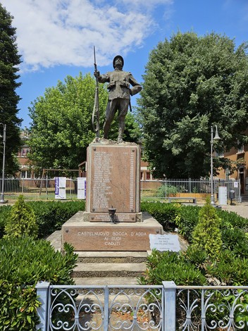 Monumento ai Caduti di Castelnuovo Bocca d'Adda, con statua e scritte commemorative, circondato da verde.